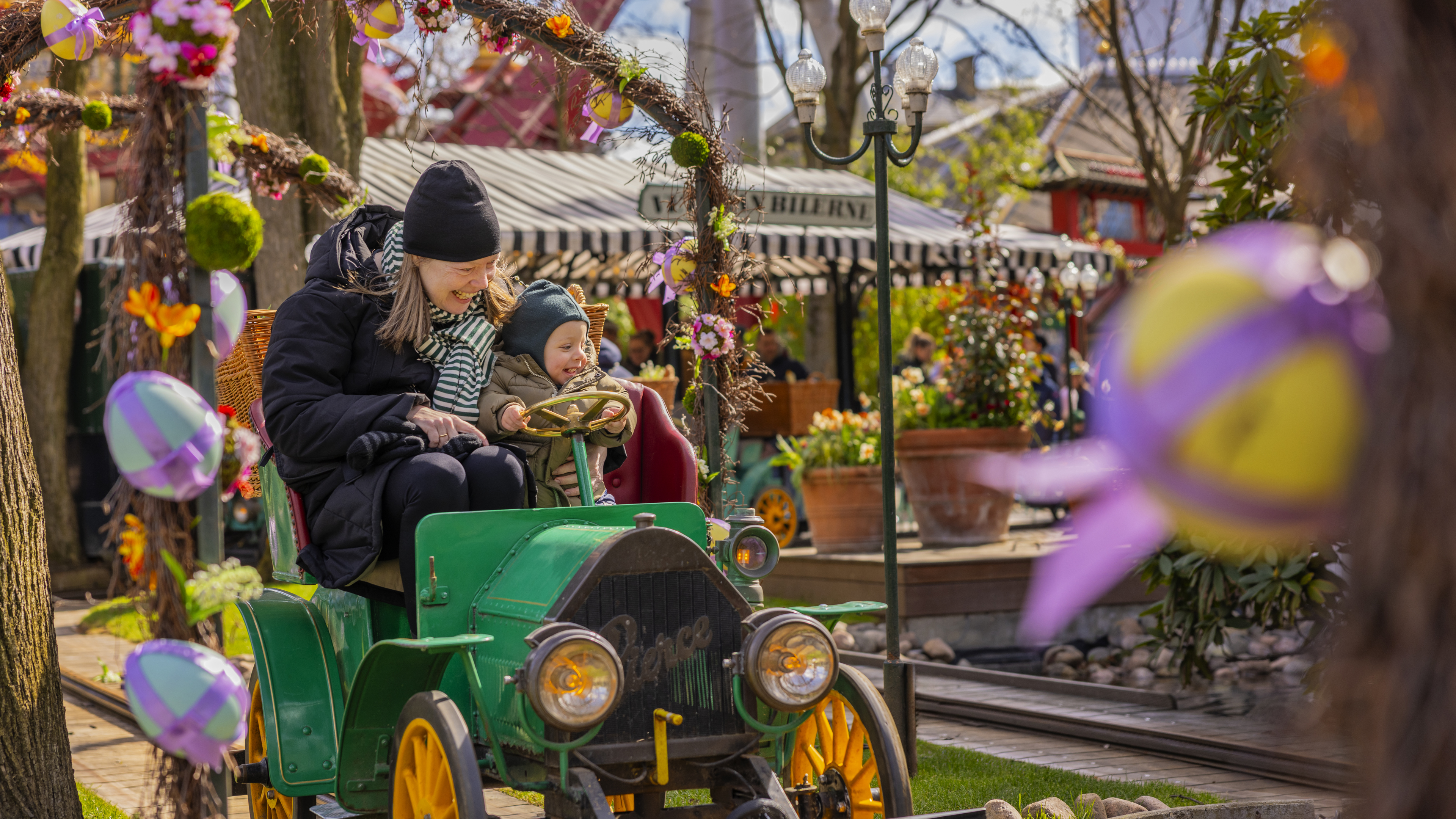 A mother and her child ride in a vintage car at Tivoli, surrounded by Easter decorations. The car is green with orange wheels, and the atmosphere is festive with colorful eggs and flowers. 