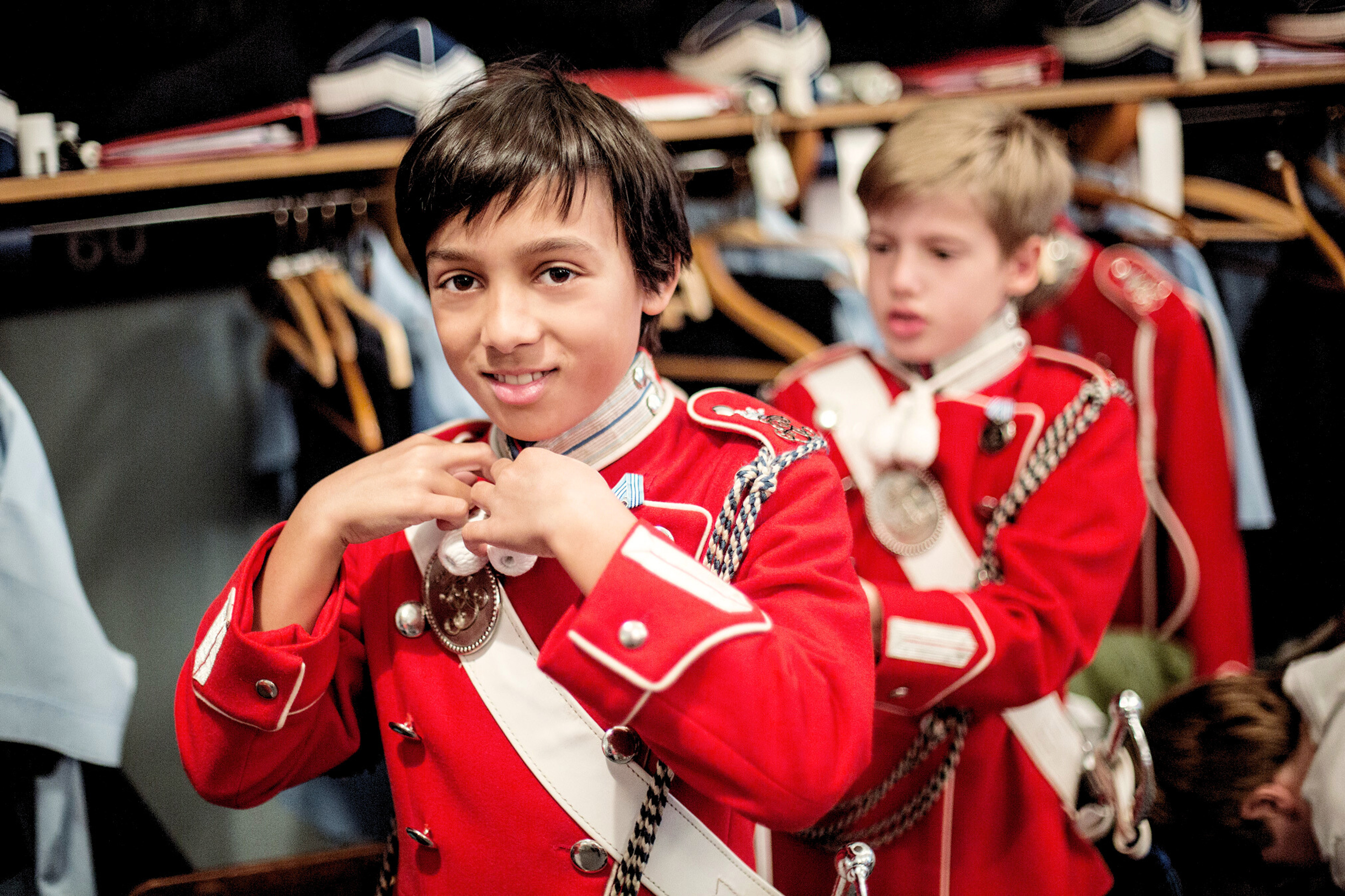 A young Tivoli guard in a red uniform is adjusting his collar, preparing for duty. In the background, another young guard, also in a red uniform, is getting ready. They are both in a dressing room with uniforms and hats hanging in the background.