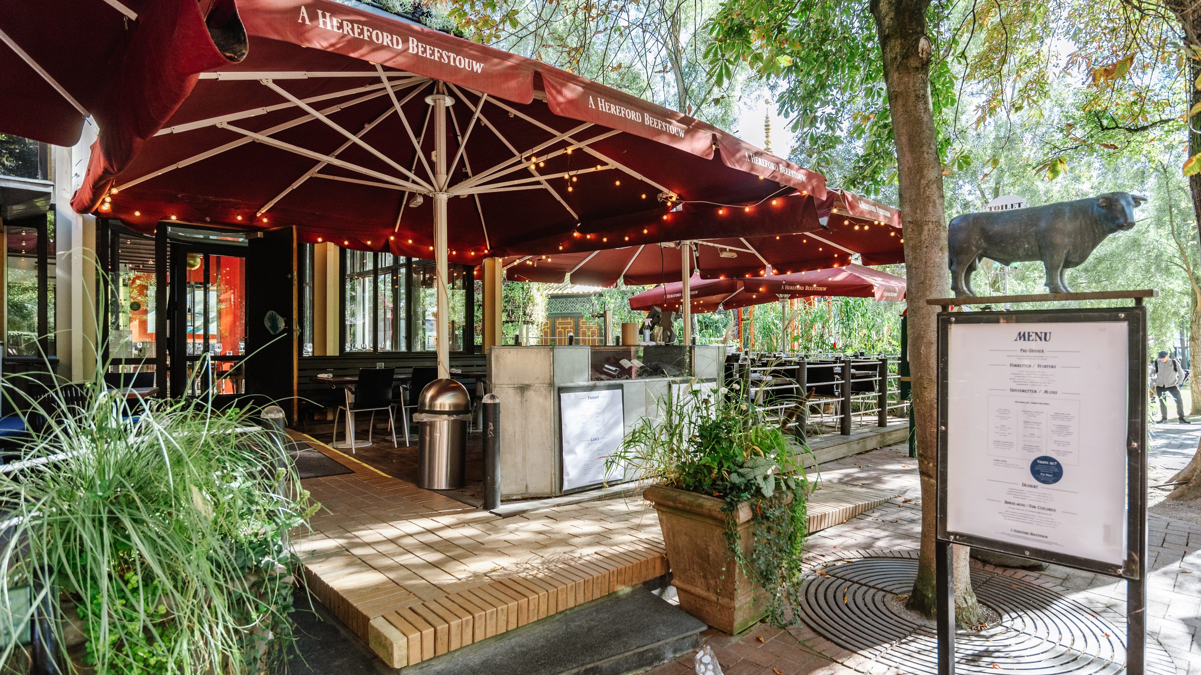 Restaurant med terasse og grønne planter med rød parasol og lamper hængende rundt i kanten af parasollen.