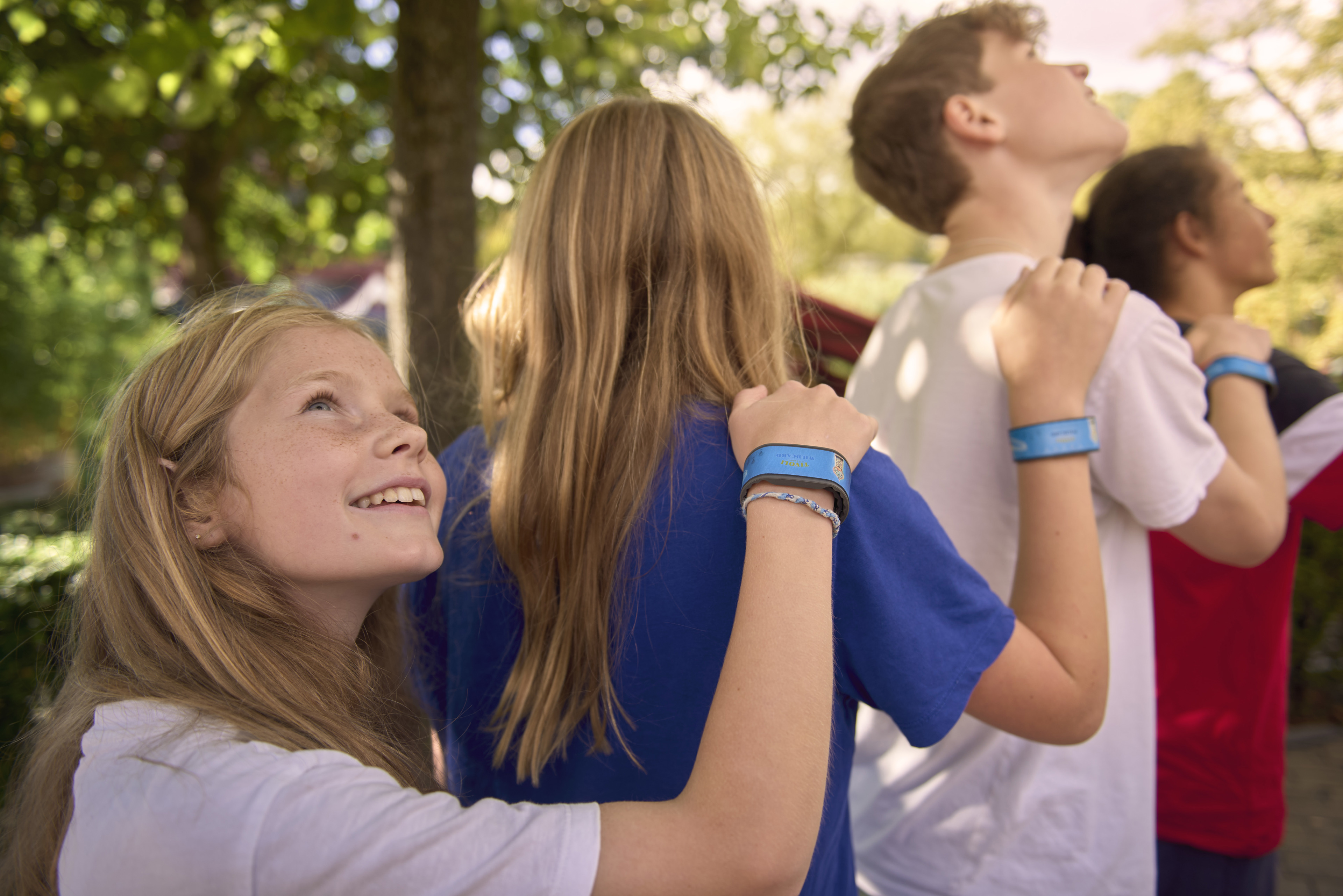 En gruppe børn står i kø i Tivoli, hvor en pige smilende ser opad, mens hun holder hånden på skulderen af barnet foran sig. De har alle turarmbånd på til forlystelserne.