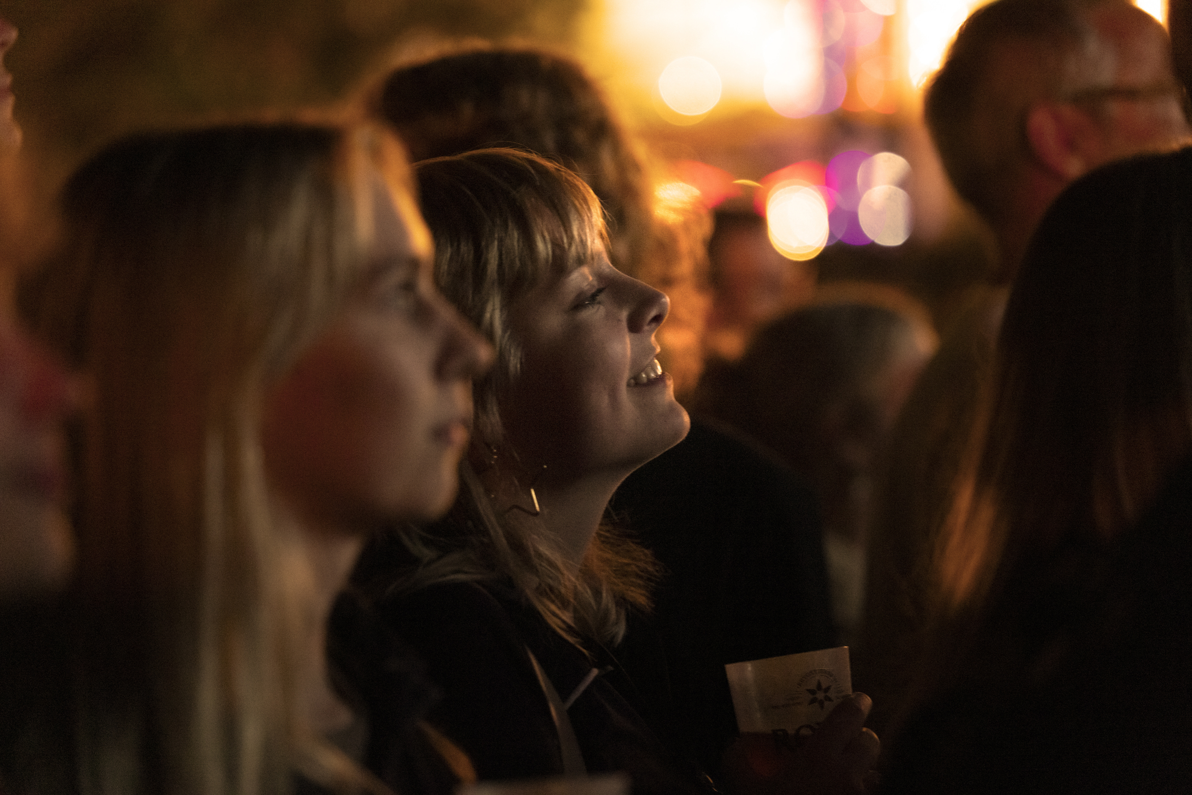 En kvinde står blandt en gruppe mennesker og ser smilende op mod scenen under en koncert i Tivoli. Baggrunden er oplyst af varme lys, der skaber en festlig atmosfære.