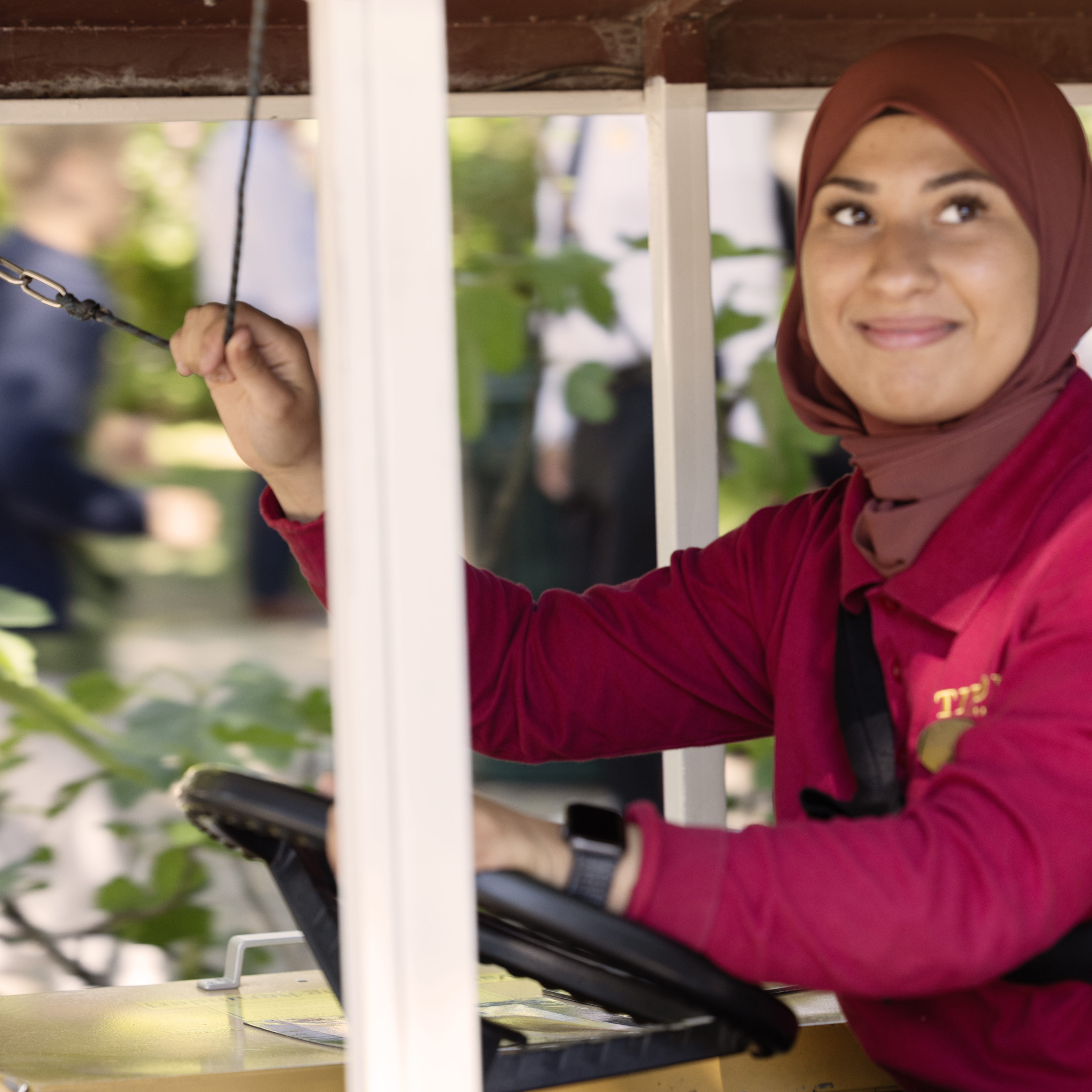 En smilende medarbejder i Tivoli, der betjener en forlystelse. Hun er iført en rød uniform og et matchende tørklæde, mens hun styrer Linje 8s gule sporvogn og kigger ud mod gæsterne.