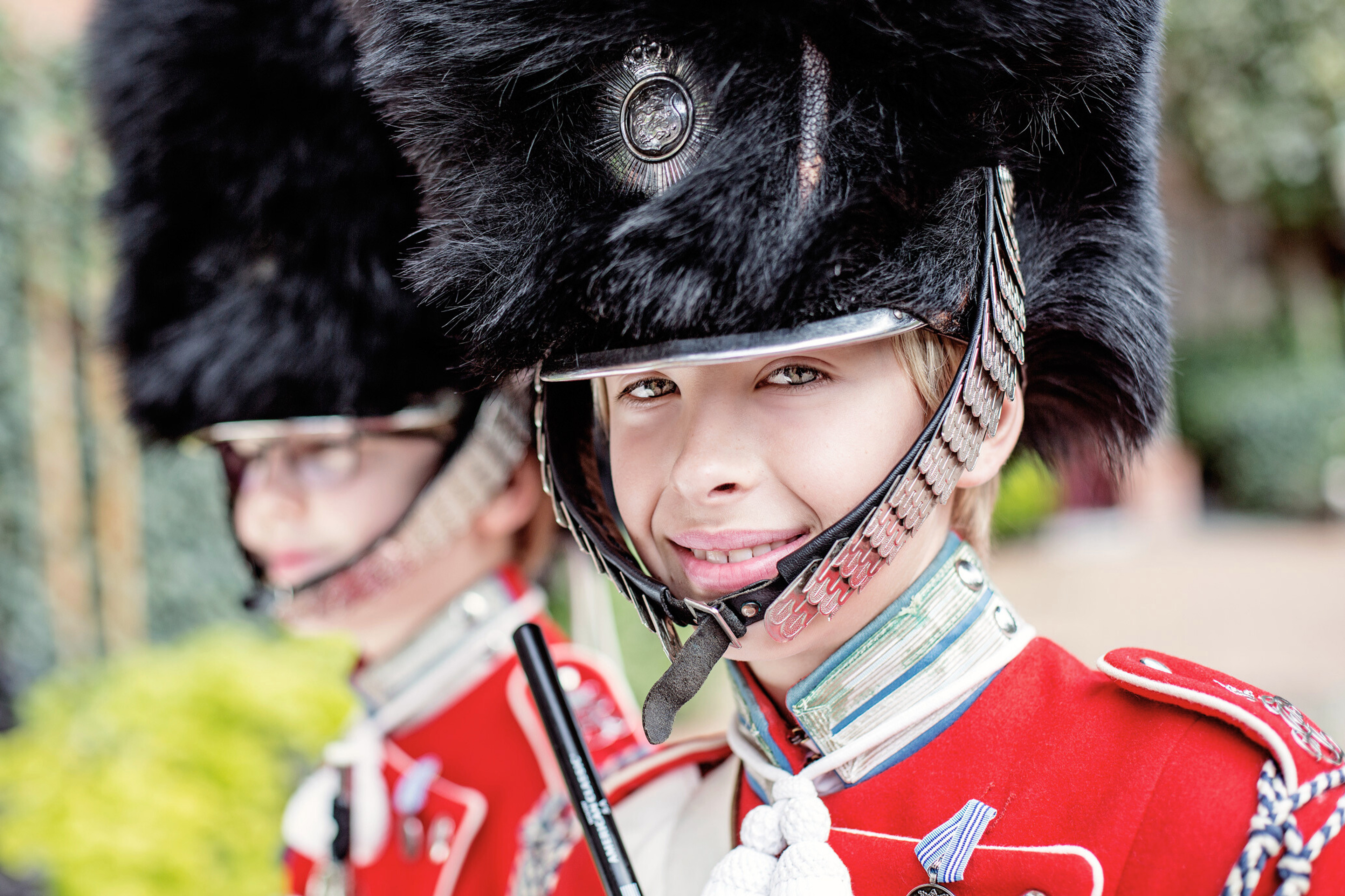 Nærbillede af en smilende dreng fra Tivoli-Garden i sin røde uniform og sorte bjørneskindshue. En anden garder er sløret i baggrunden.