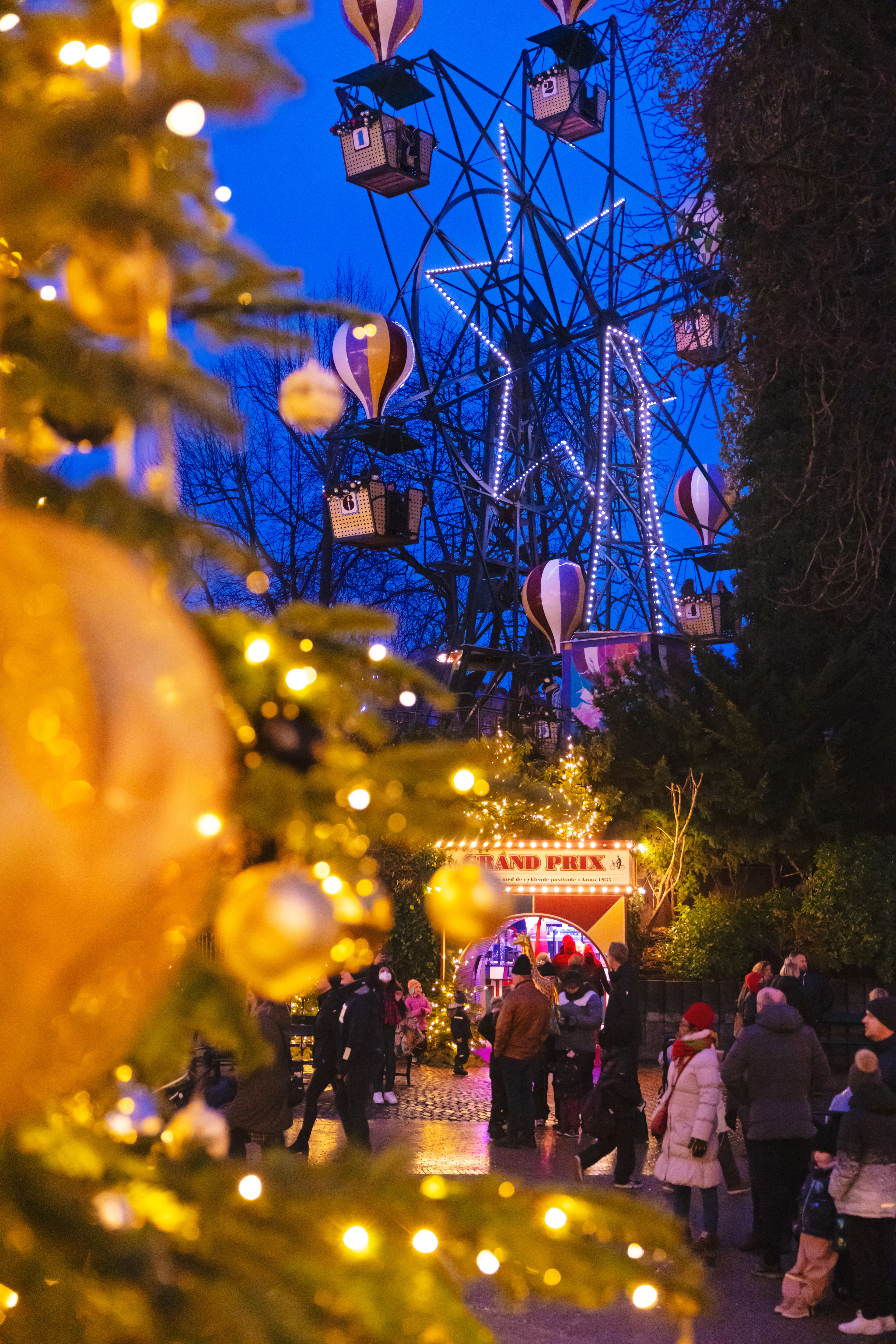 Et billede af Ballongyngen i Tivoli, oplyst af farverige julelys om aftenen. I forgrunden ses en gylden julekugle og et juletræ med lys, mens besøgende går rundt foran Grand Prix.