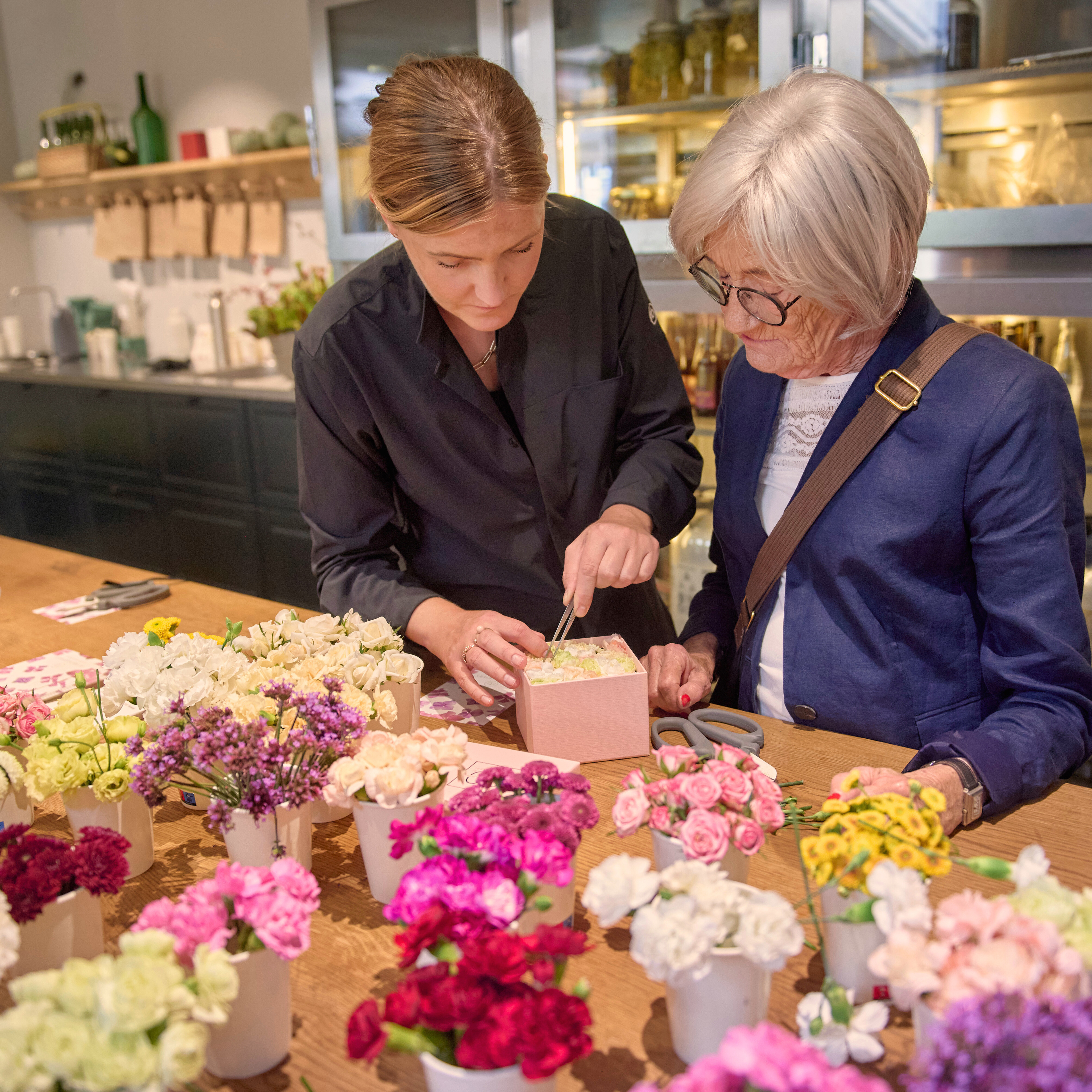 En medarbejder hjælper en kvinde med at pakke blomster ind i en butik i Tivoli. Bordet er fyldt med farverige blomster, og butikken har en varm og indbydende atmosfære.