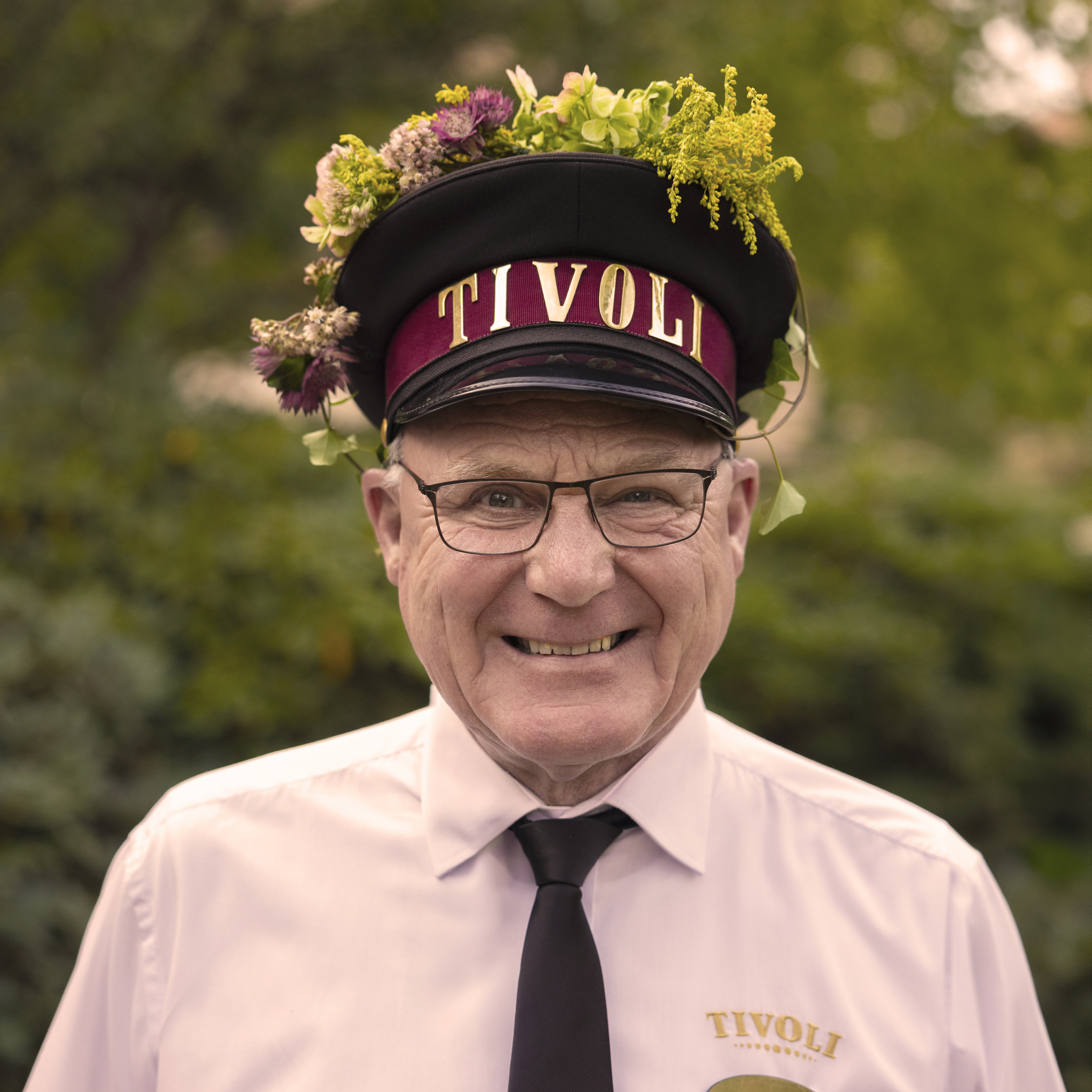 En mandlig medarbejder iført en Tivoli-uniform og en Tivoli-hat. Han smiler og bærer blomsterdekorationer på sin hat. Han står udenfor med en grøn baggrund. 