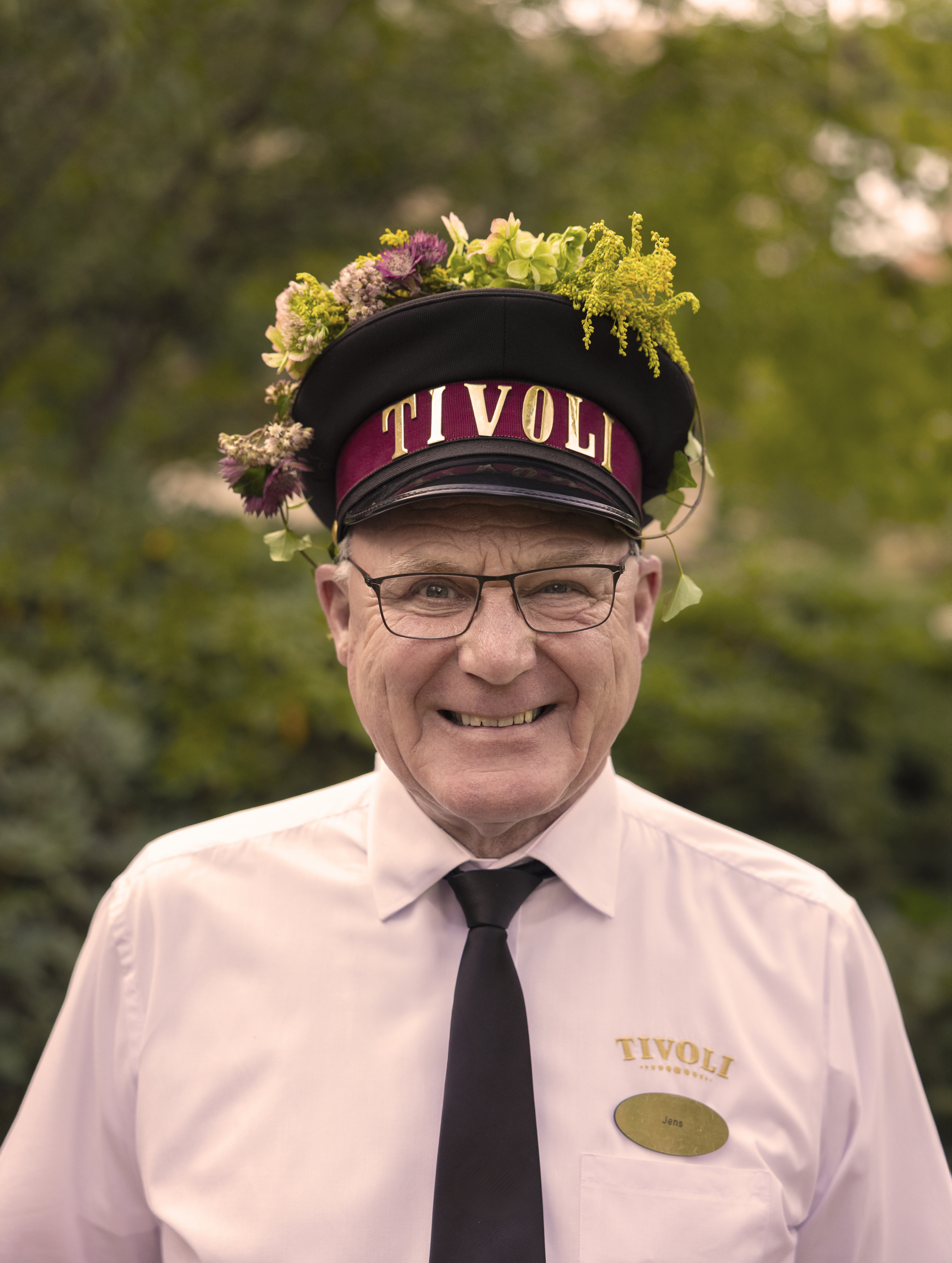 A male employee wearing a Tivoli uniform and a Tivoli hat. He is smiling and has floral decorations on his hat. He is standing outside with a green background.