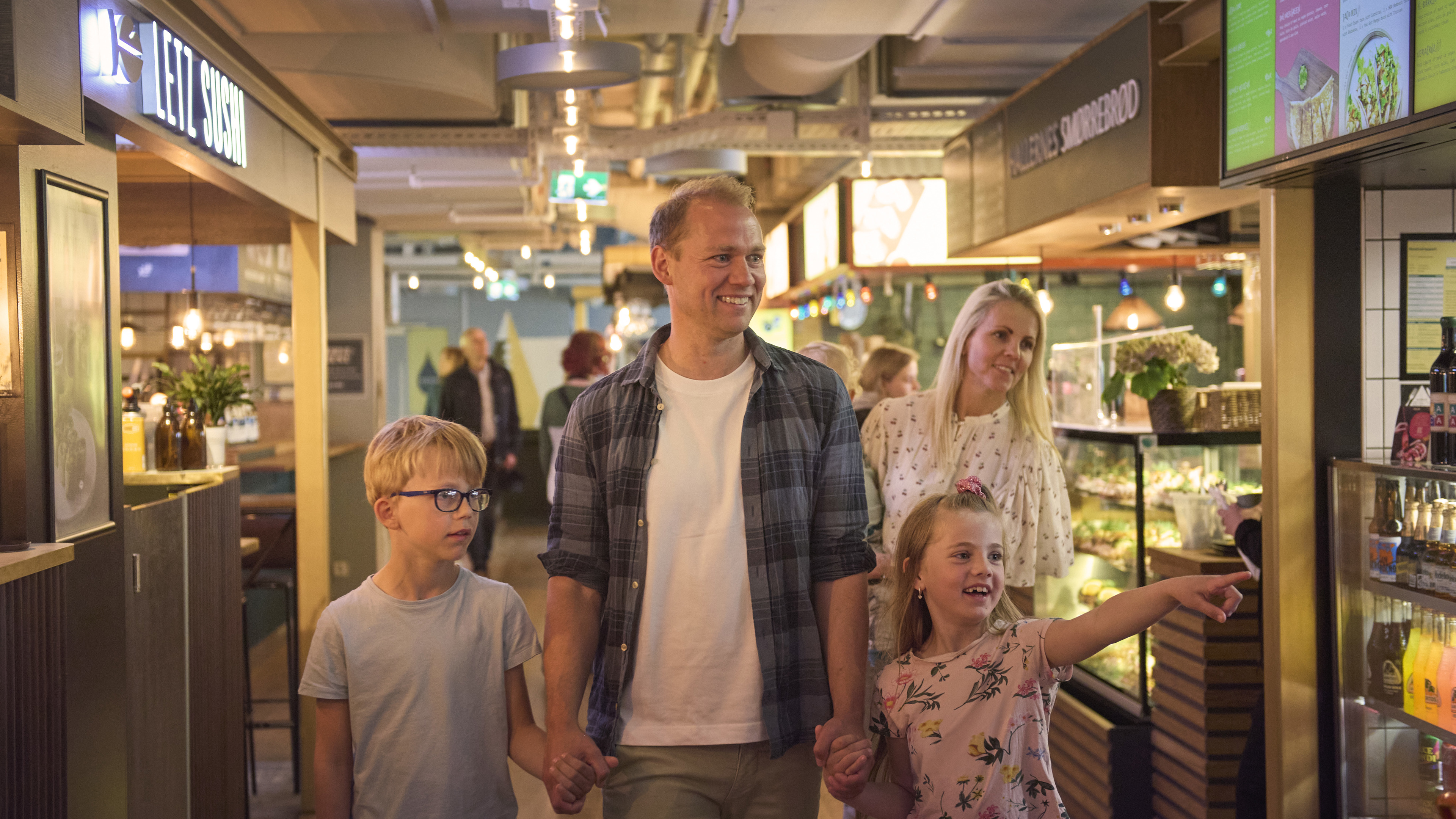 En familie på fem går blandt boderne i Tivoli Food Hall. Faren går forrest med sin datter i den ene hånd og søn i den anden. Bag dem går mor med et barn på hoften. De kigger interesseret på en bod udenfor billedet. 