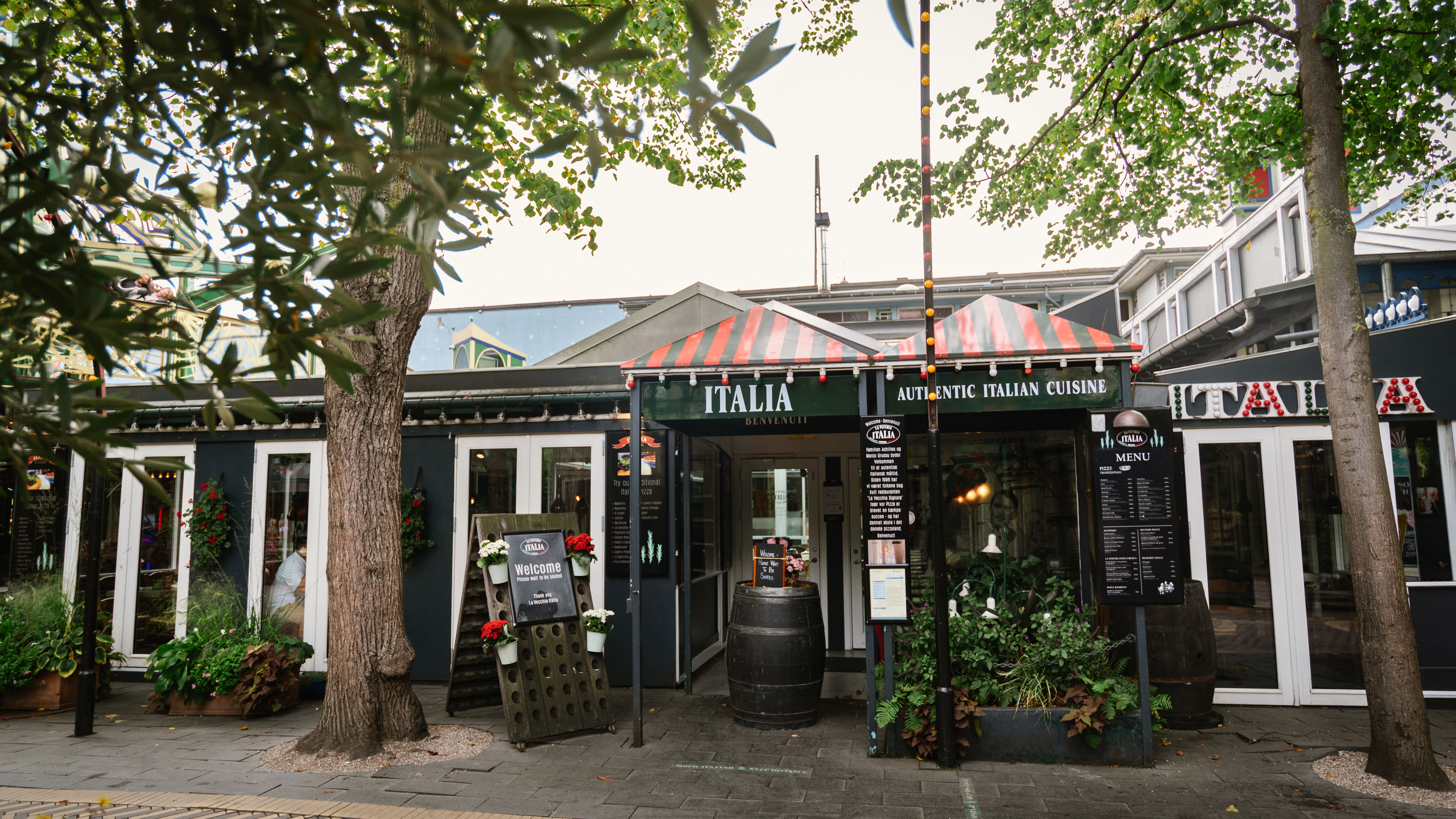 Restaurant med grønne og røde markiser og et skilt med 'Italia'. Grønne træer og planter rundt om.