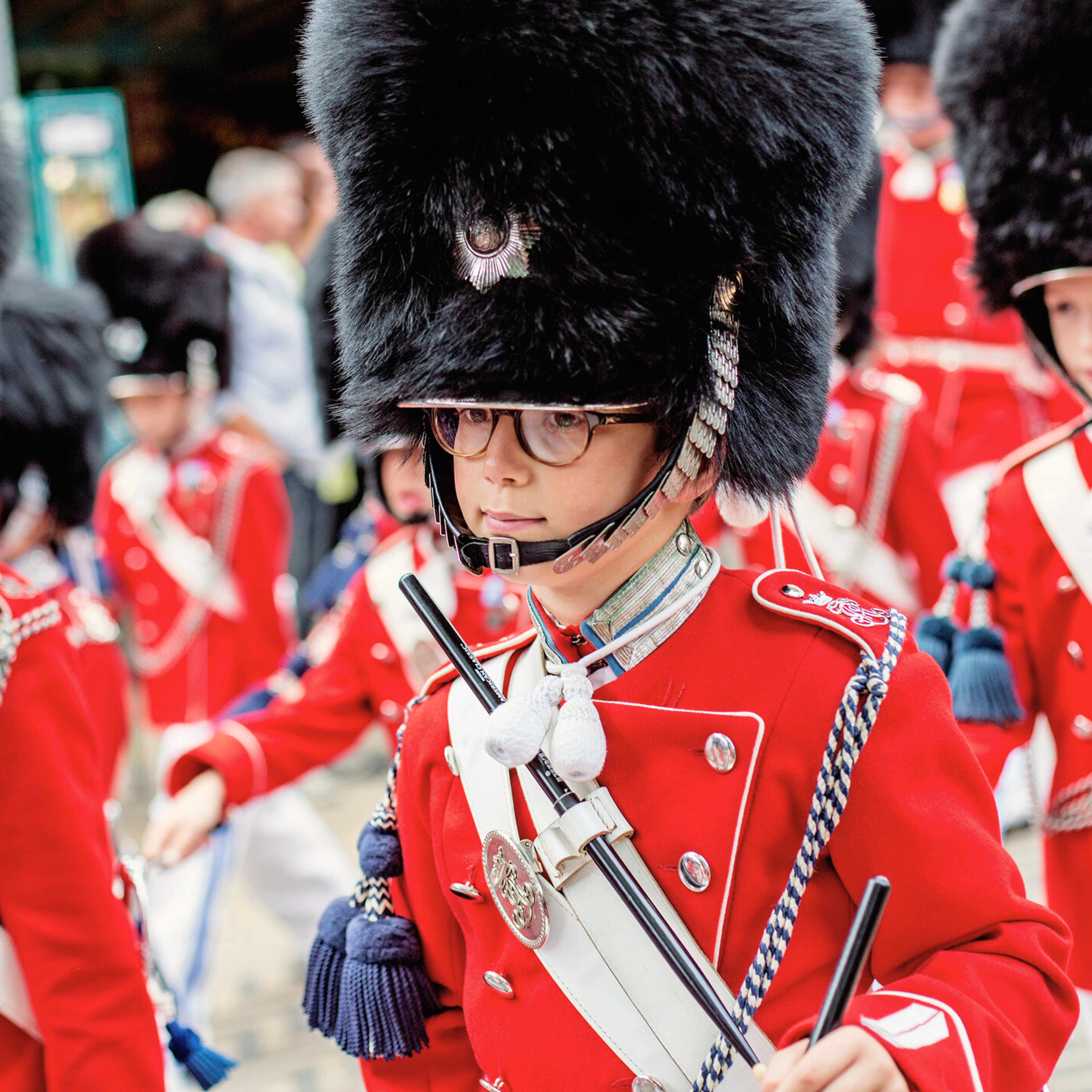 En Tivoli-gardist iført en rød uniform og den karakteristiske høje bjørneskindshue står i fokus. Han bærer briller og holder på sin trommestik, klar til optræden. I baggrunden ses flere andre gardister i formation.