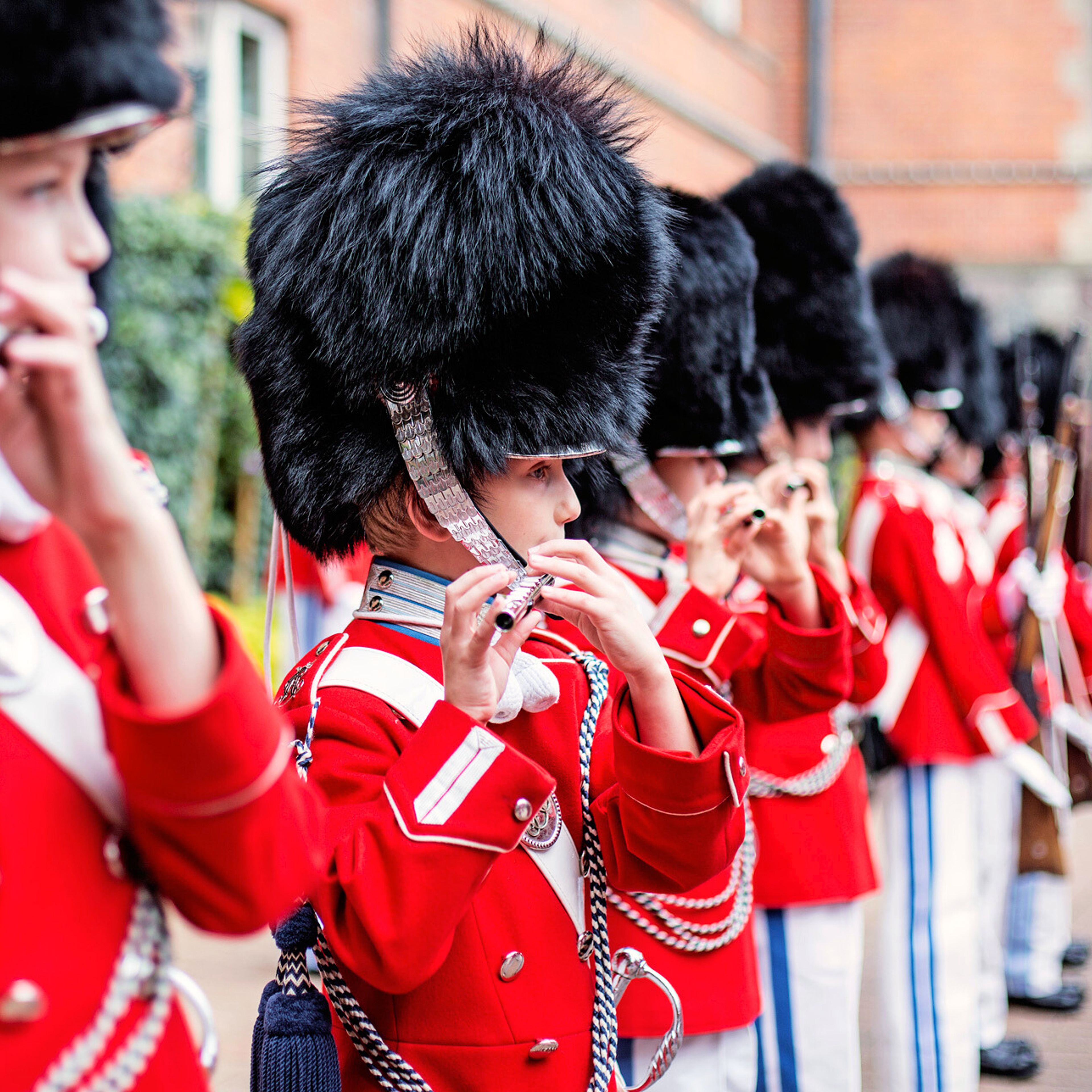 En række medlemmer af Tivoli-Garden i røde uniformer og bjørneskindshuer spiller deres fløjter i perfekt harmoni under en parade, fuldt fokuserede på musikken.