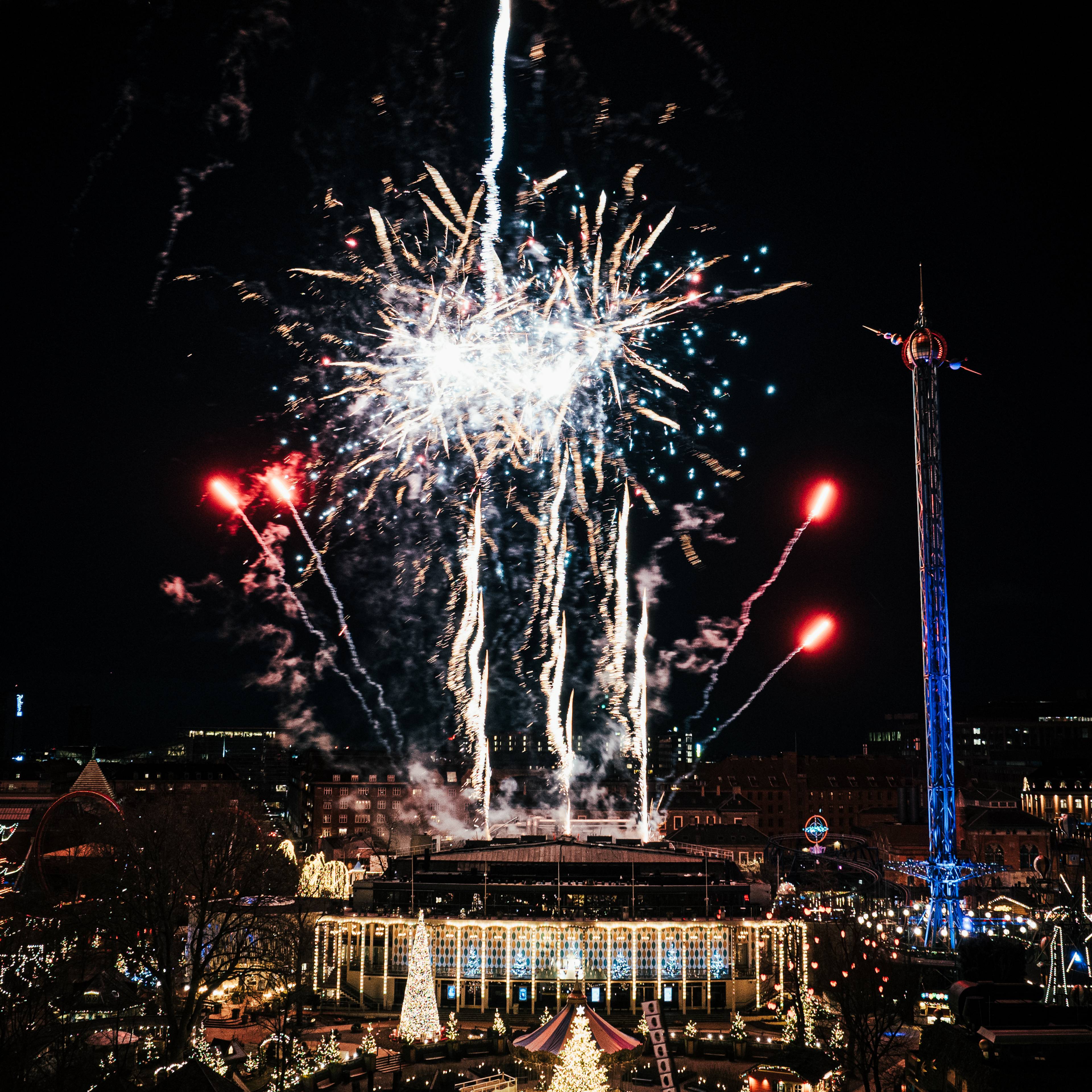 Et spektakulært fyrværkerishow lyser nattehimlen op over Tivoli. Julebelysning og forlystelser i baggrunden skaber en festlig stemning.