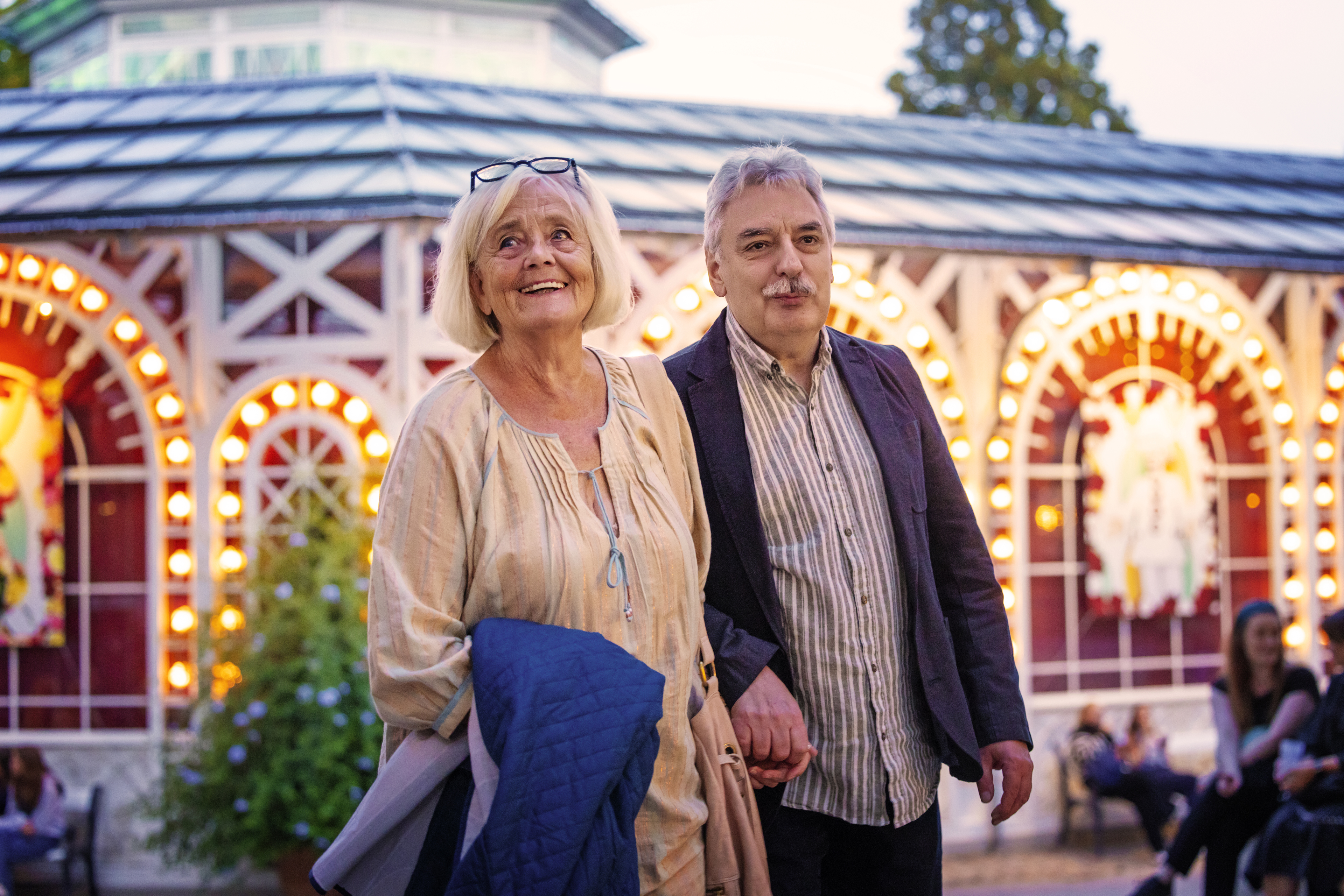 An elderly couple is seen smiling and standing in front of the Glass Hall, illuminated by lanterns and lights in the evening.