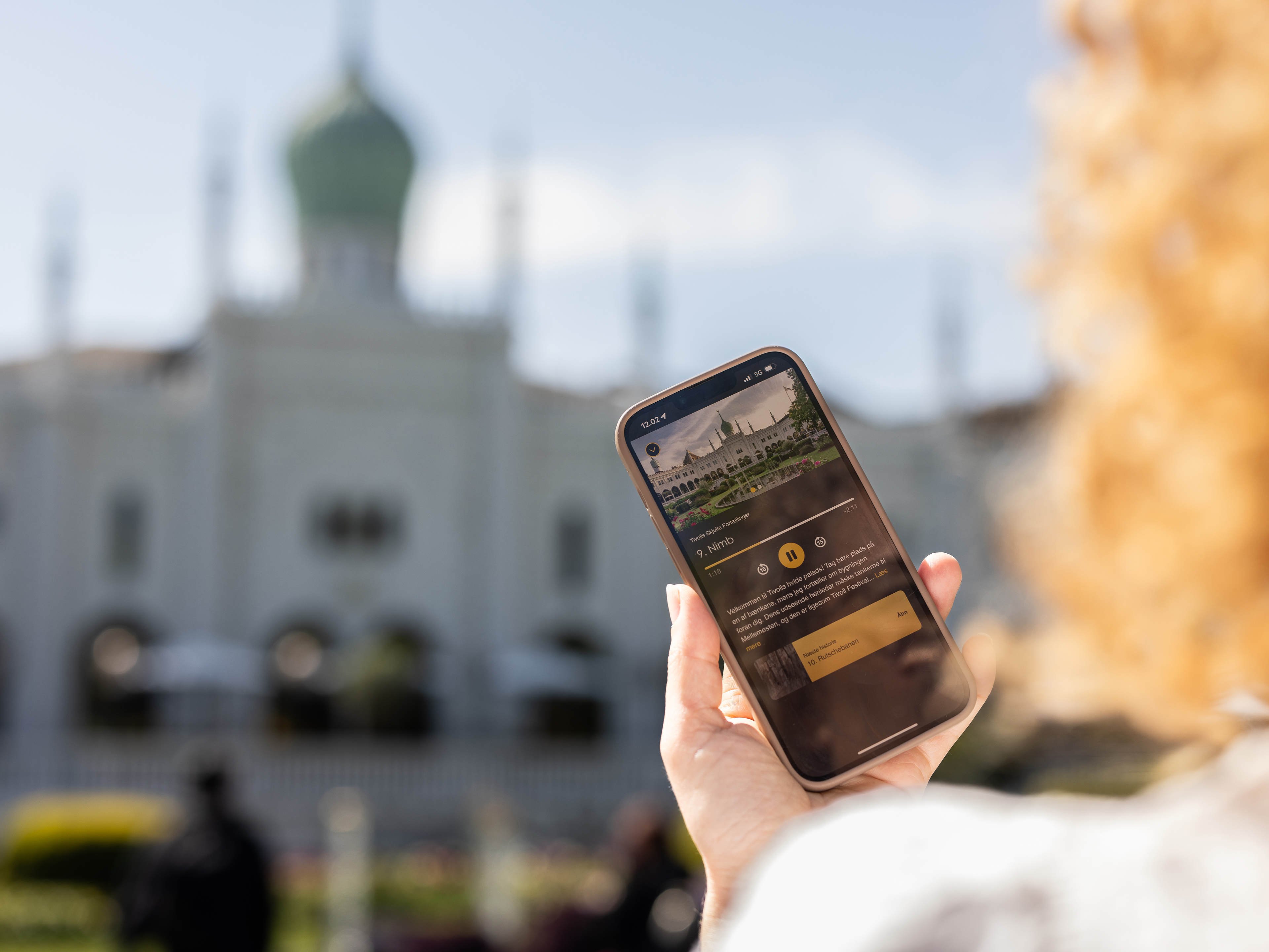 En person holder en smartphone, der viser information om Tivoli, med den ikoniske bygning Nimb i baggrunden på en solrig dag.